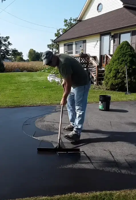 Worker applying driveway sealer on a driveway with a squeegee, near a suburban house