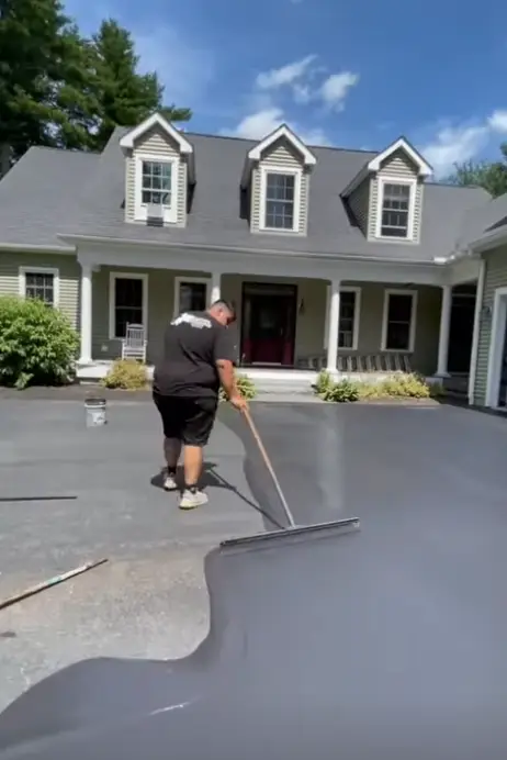 A person applying driveway sealer in front of a suburban house on a sunny day