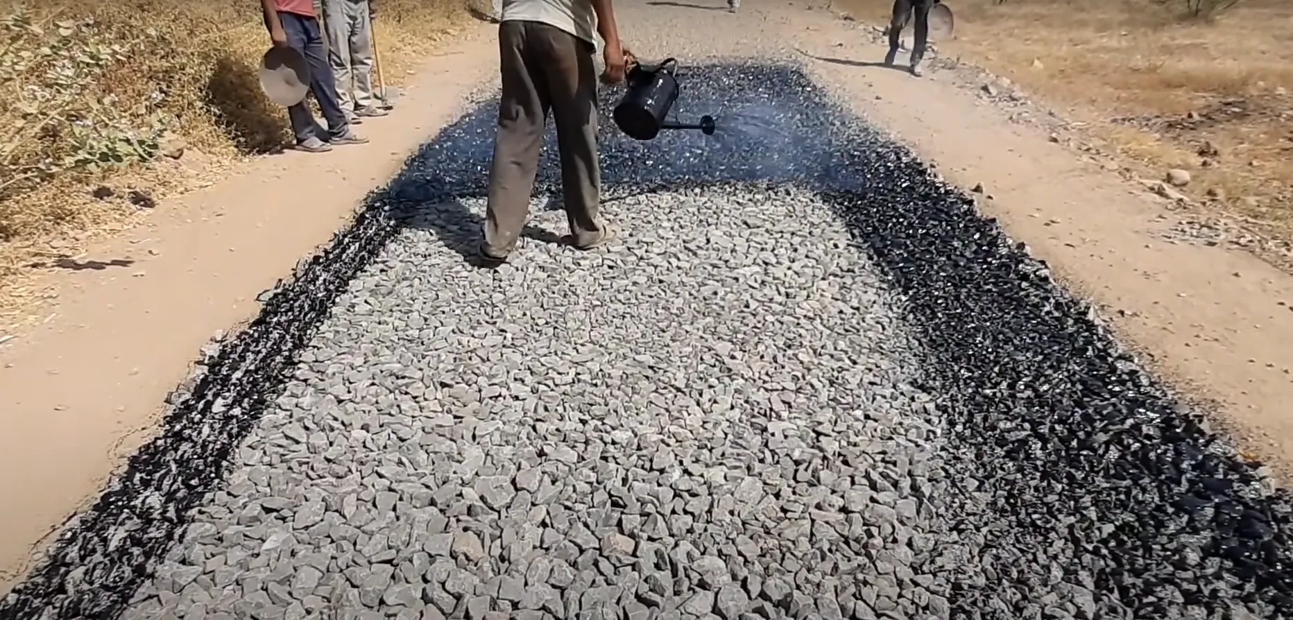 Worker applying bitumen on a layer of crushed stone for macadam road construction.