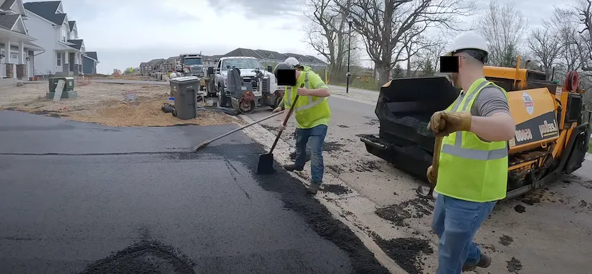 Two workers laying and spreading asphalt on a residential street with a paving machine in the background.