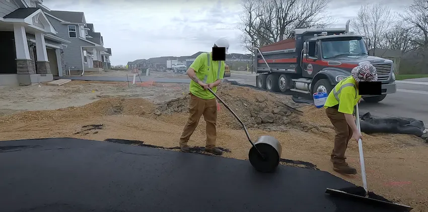 Two workers compacting freshly laid asphalt with a roller and a shovel on a construction site.