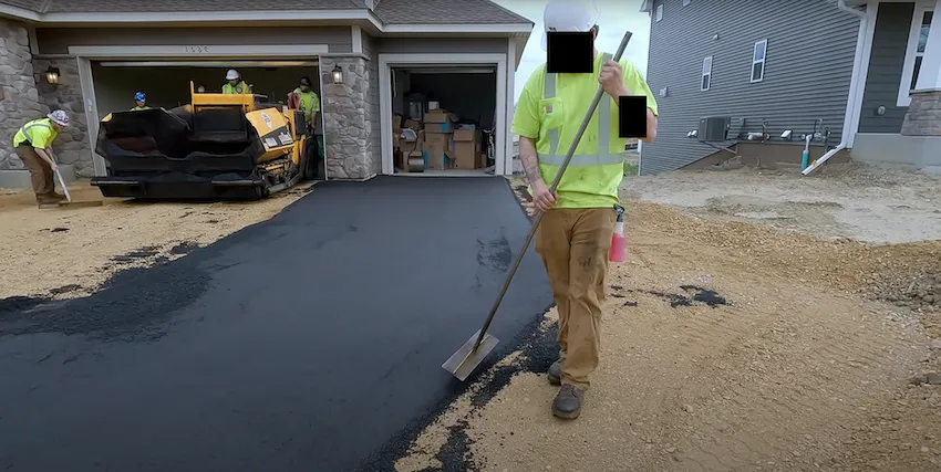 Worker smoothing asphalt on a residential driveway with a paving machine in the garage.