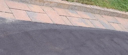 A close-up view of a driveway with a combination of asphalt and interlocking stone pavers, showing a clean transition between the two materials and bordered by a stone curb.