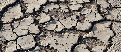 Image en gros plan d'une surface de service au volant dégradée avec des fissures visibles, des nids-de-poule et des signes d'usure, tels que de la peinture décolorée et des textures rugueuses, exprimant une impression de vieillissement et de négligence.