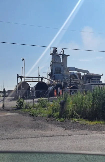 Une usine d'asphalte avec de grands réservoirs de stockage, des canalisations et une tour centrale est visible sous un ciel bleu clair.