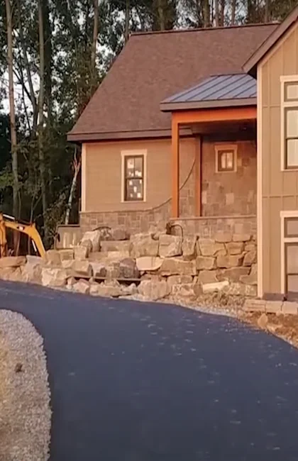 Une allée en asphalte foncé fraîchement pavée serpente devant une maison rustique et un muret de pierre paysager.