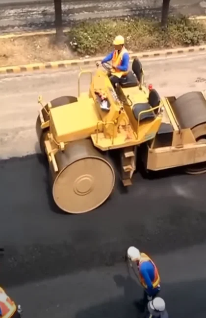 Des ouvriers du bâtiment observent un grand rouleau compresseur jaune compacter une nouvelle couche d'asphalte sur une route.